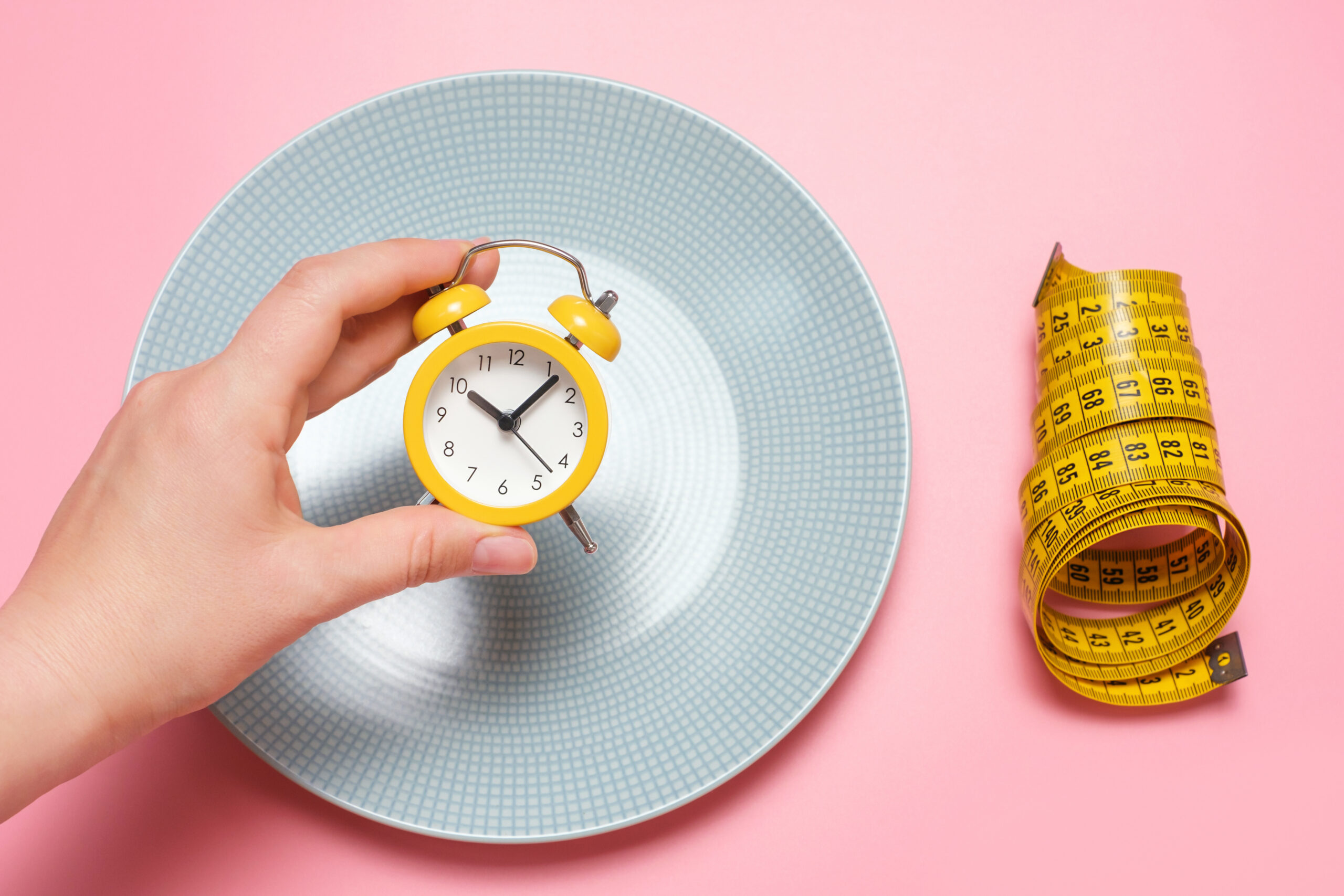 Female hand holding an alarm clock on the background of an empty plate, concept of intermittent fasting. 5-Day Fasting Diet Meal Plan